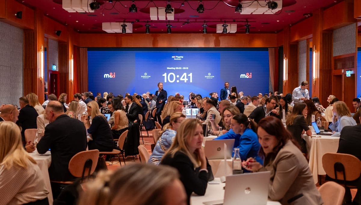 Buyers and suppliers in one-to-one meetings on the M&I event floor, the meeting countdown clock on the stage screen behind them